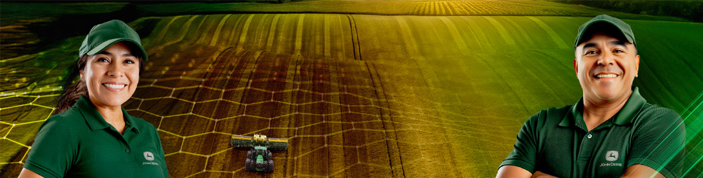 Una mujer y un hombre, ambos vistiendo gorra y camisa verdes, en un campo con una máquina John Deere al fondo.