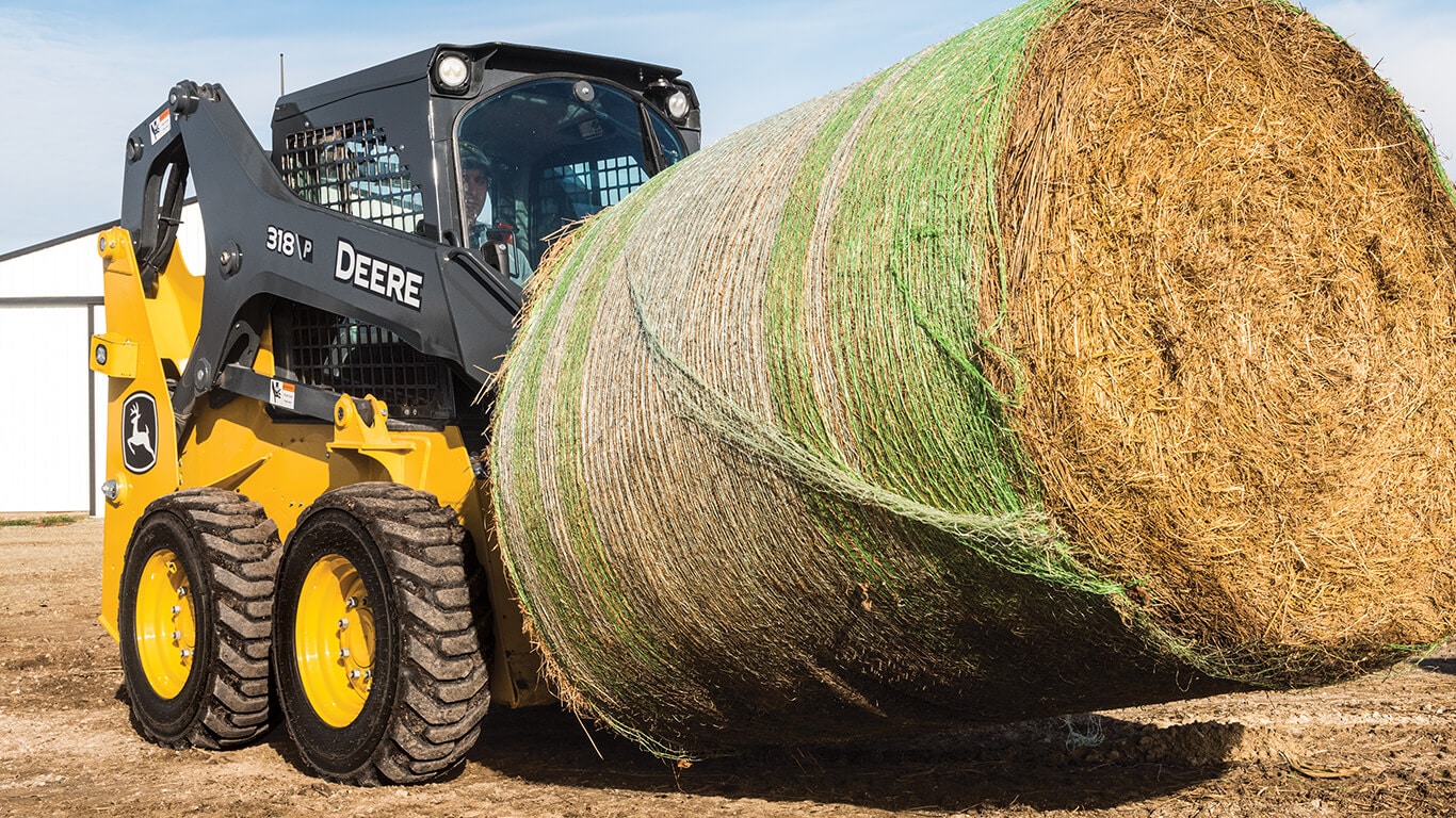 Minicargador deslizante 318 versión P que levanta y mueve fardo de heno con accesorio de horquilla Deere.