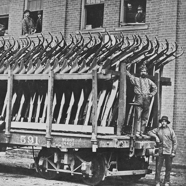 Fotografía de 1882 de tres hombres parados junto al vagón del tren de Deere & Co. cargado con arados de acero listos para ser embarcados; se ven también unos empleados de la fábrica mirando por la ventana del edificio de atrás.