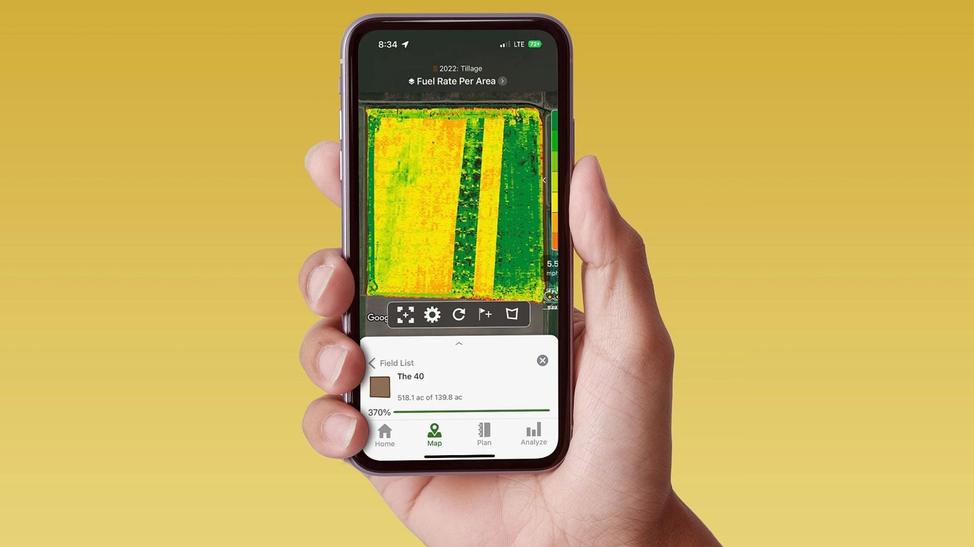 Una mano sostiene un teléfono inteligente que muestra el John Deere Operations Center con datos de caudal de combustible.