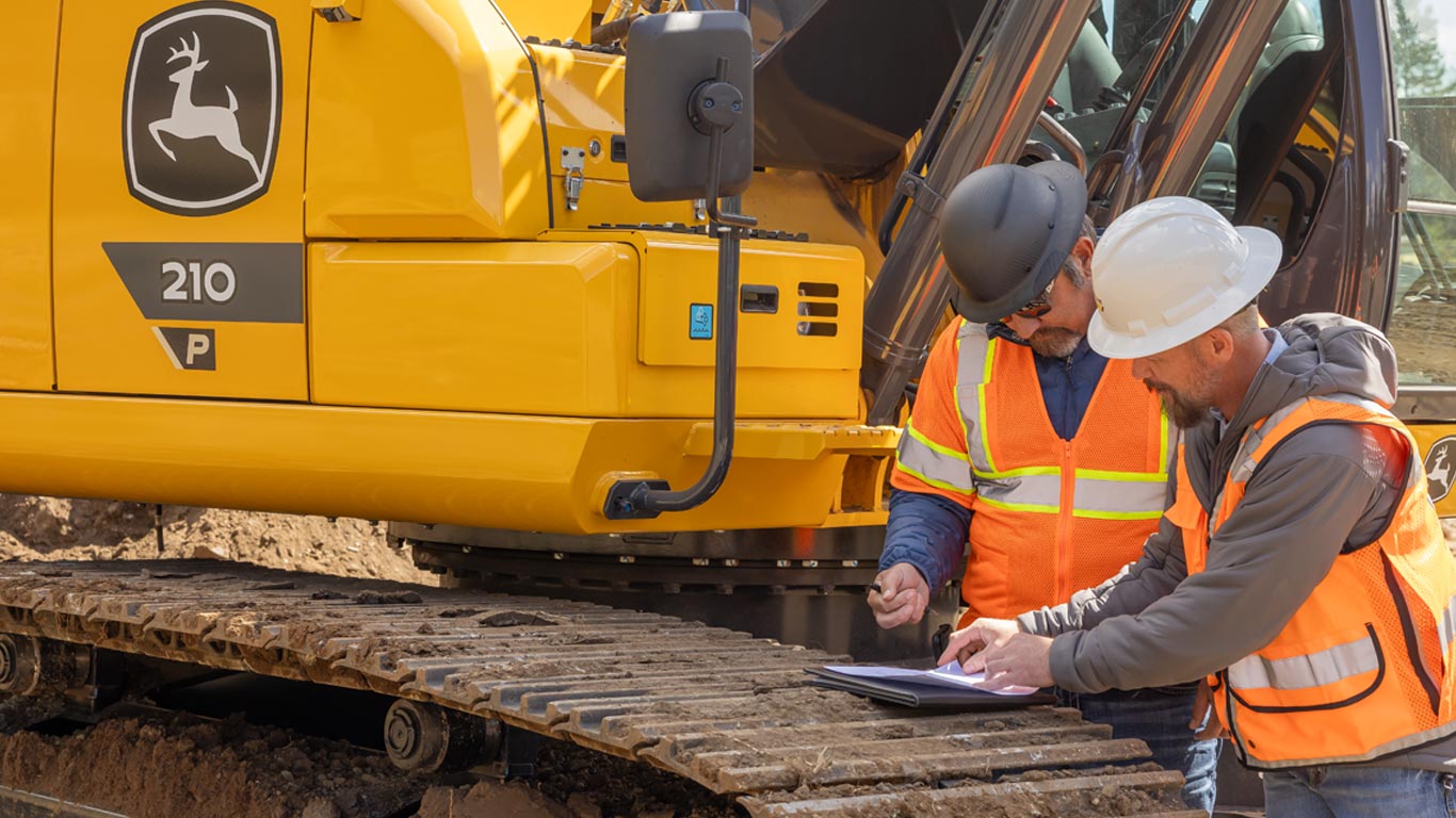 Dos trabajadores de la construcción con equipo de seguridad revisan documentos en una carpeta al lado de una excavadora amarilla.