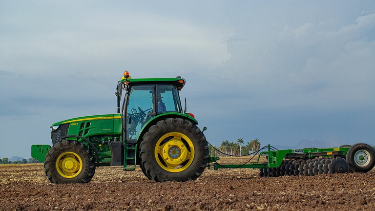 Tractor 6125E visto de lado trabajando la tierra con arado de disco. El tractor es manejado por un hombre en camisa azul de cuadros y sombrero de paja.