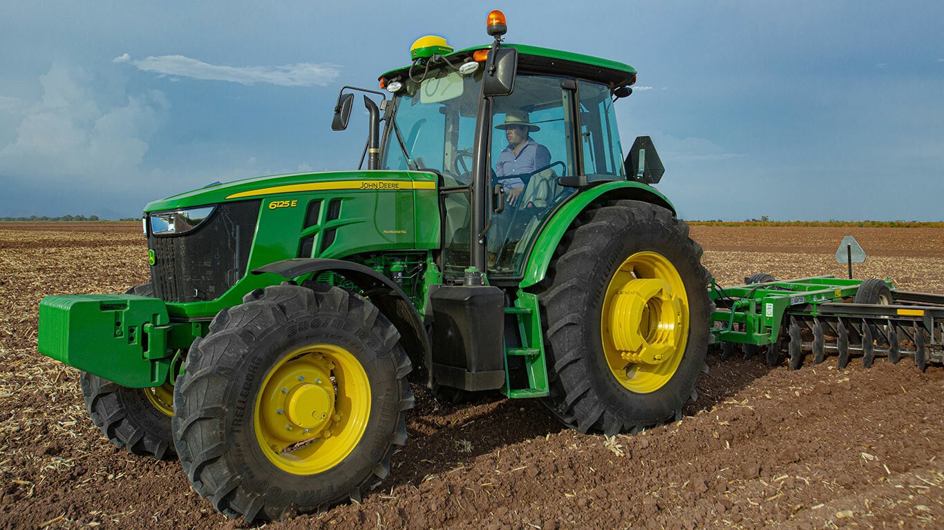 Tractor 6125E visto en diagonal trabajando la tierra con arado de disco. El tractor es manejado por un hombre en camisa azul de cuadros y sombrero de paja.