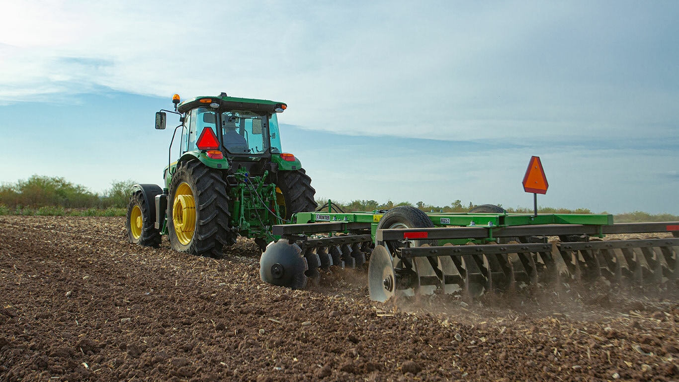 Tractor 6125E visto de atrás trabajando la tierra con arado de disco. El tractor es manejado por un hombre en camisa azul de cuadros y sombrero de paja.