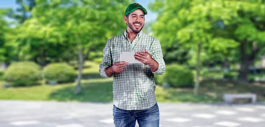 Hombre jóven con gorra verde John Deere, camisa de cuadros y pantalón de mezclilla. En las manos tiene un iPad y está en un parque.