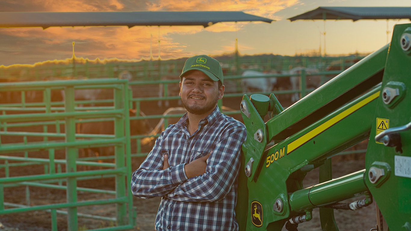 Jóven en camisa de cuadros y gorra John&nbsp;Deere, recargado en cargador frontal 500M con los brazos cruzados. De fondo está el atardecer y ganado en su establo.