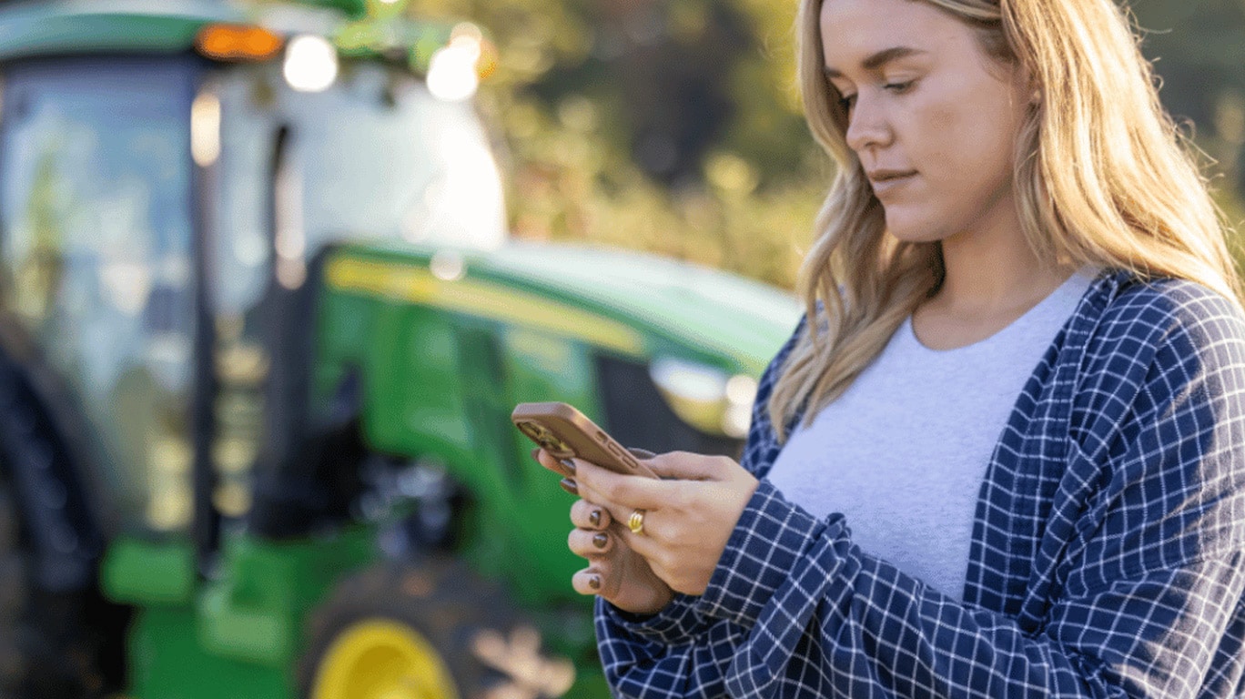 Uma chica blanca, rubia, usando su celular frente a una máquina John Deere