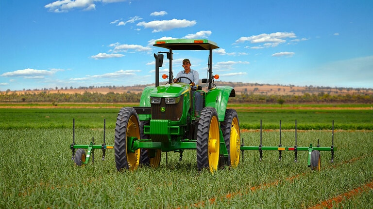 tractor 6105E sin cabina con techo trabajando el campo con arado visto de frente con conductor adentro vestido en camisa clara y gorra John Deere