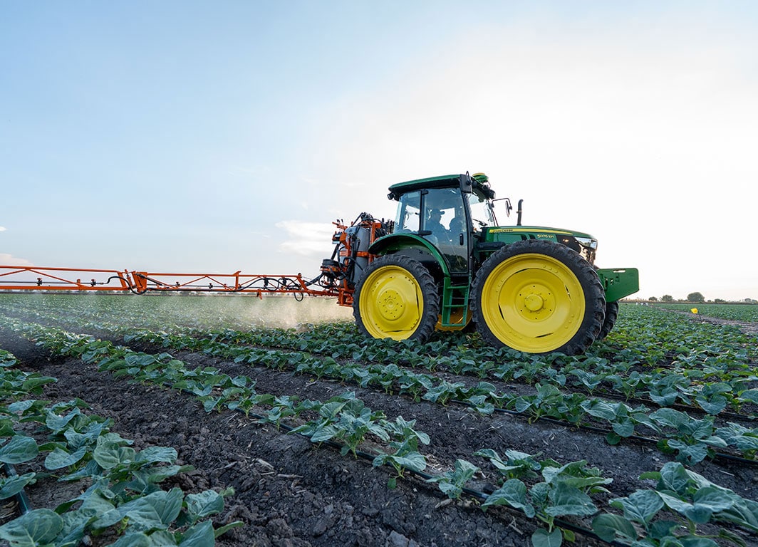 Tractor John Deere aplicando insumos en un cultivo, avanzando entre hileras en un campo abierto al atardecer.
