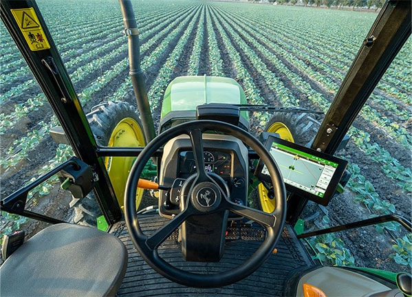 Vista desde la cabina de un tractor John Deere alineado con hileras de cultivo en un campo abierto.