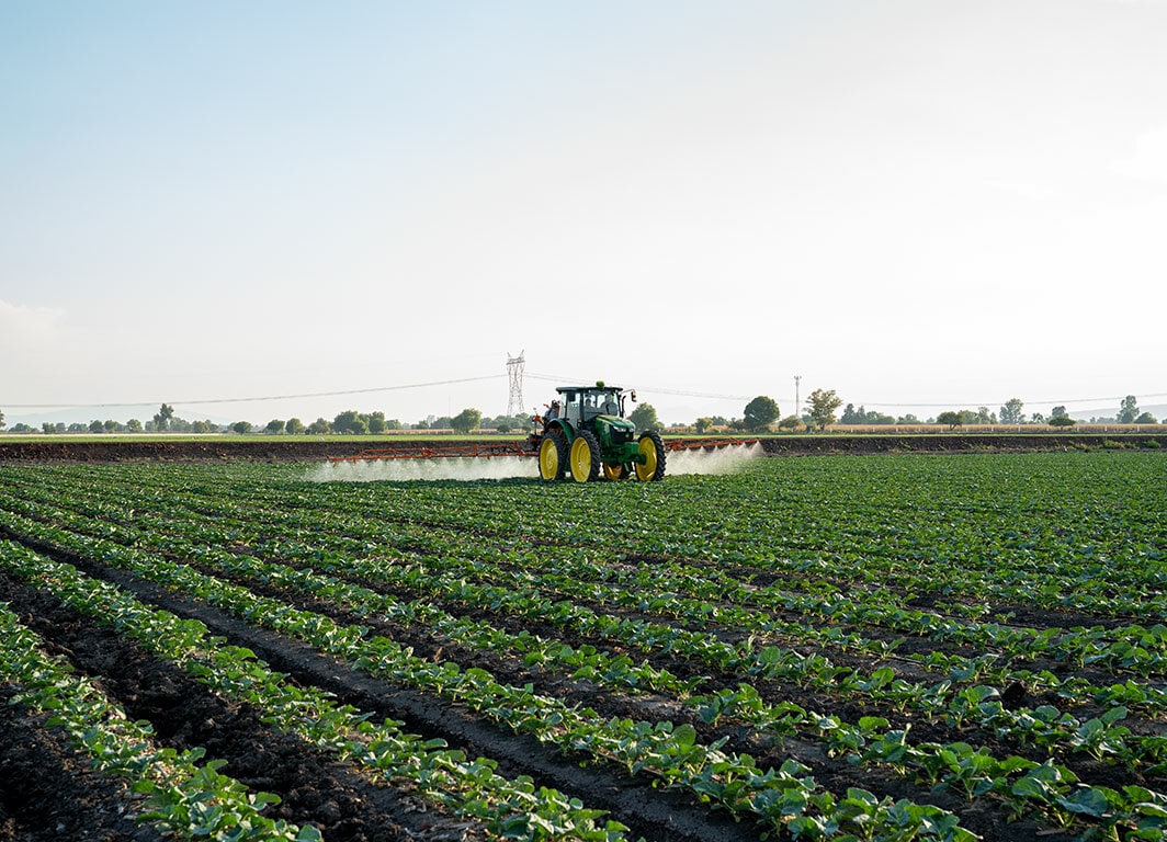 Tractor aplicando insumos en un cultivo, avanzando entre hileras en un campo abierto al atardecer.
