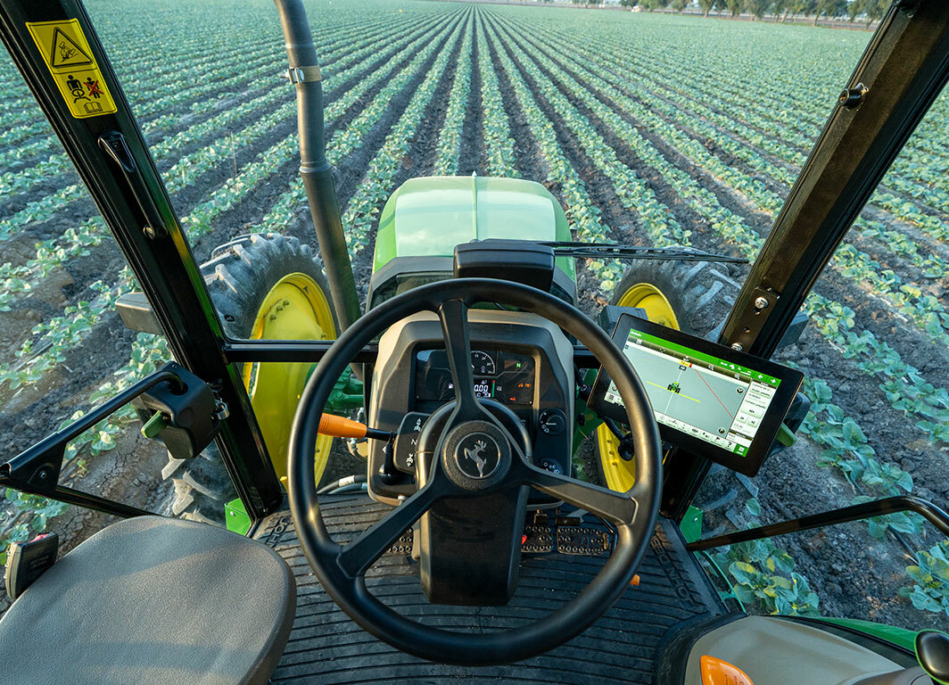 Vista desde la cabina de un tractor John Deere alineado con hileras de cultivo en un campo abierto.