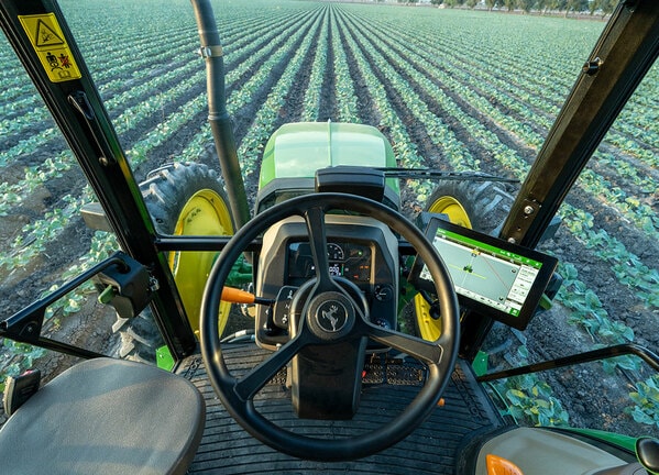 Vista desde la cabina de un tractor John Deere alineado con hileras de cultivo en un campo abierto.