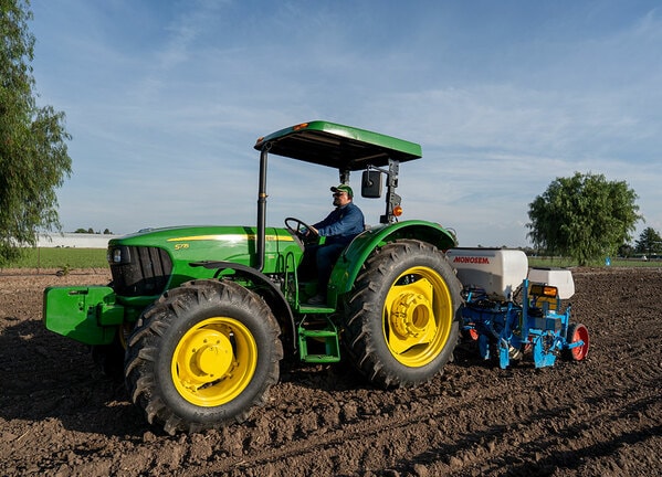Tractor John Deere trabajando en un campo con sembradora Monosem, avanzando sobre suelo preparado al atardecer.