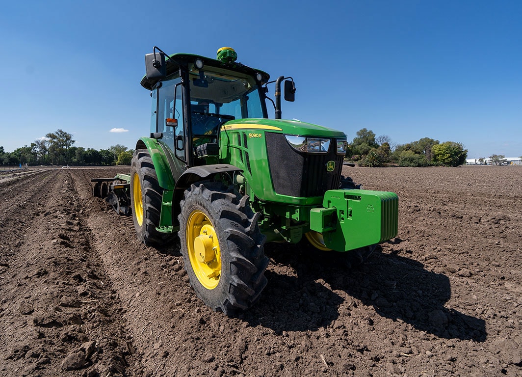 Tractor John Deere haciendo labranza, avanzando entre hileras en un campo abierto en el día con cielo azul.
