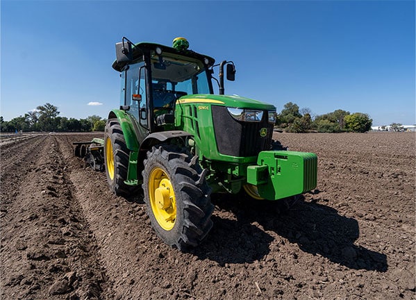 Tractor John Deere haciendo labranza, avanzando entre hileras en un campo abierto en el día con cielo azul.