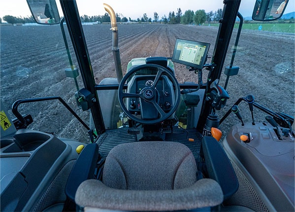 Vista desde la cabina de un tractor John Deere alineado con hileras de cultivo en un campo preparado.