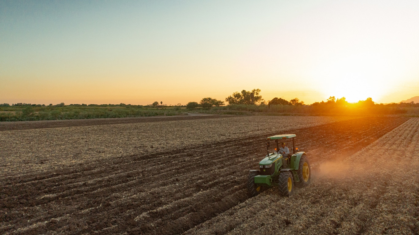 Fotografía tomada con dron de tractor 6120E trabajando la tierra con arado con la puesta del sol de fondo.